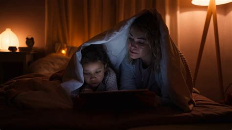 Happy Mother With Her Little Daughter Lying Under Blanket And Watching Movie On Tablet At Home