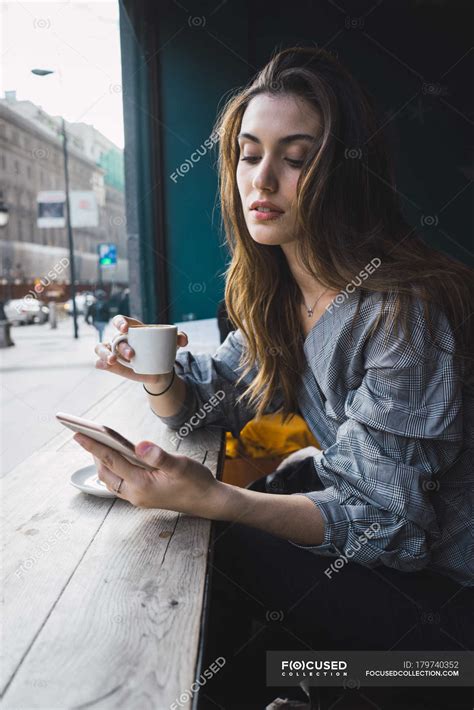 portrait of brunette girl drinking coffee at cafe and browsing smartphone — indoors woman