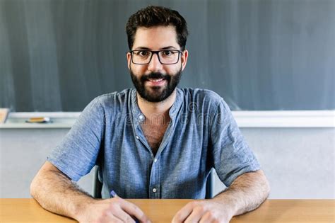 Cheerful Portrait Of Male Teacher Sitting At Desk In Classroom Stock