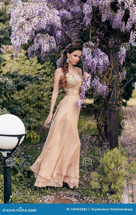 Beautiful Sensual Woman With Dark Hair In Elegant Dress Posing In Garden With Flowering Wisteria