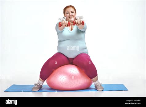 Red Haired Chubby Woman Is Sitting On The Ball And Keep The Dumbbells