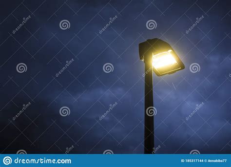 Street Lamp Lit At Twilight With Trees In The Background And Rain