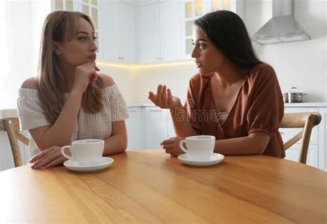 Young Women Talking While Drinking Tea At Table In Kitchen Stock Photo