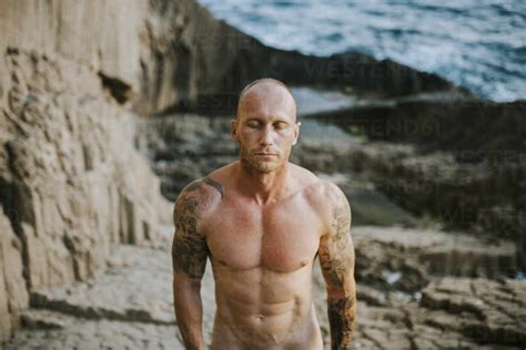 Tattooed Nudist Standing On Volcanic Rocks By The Sea With Closed Eyes Stock Photo