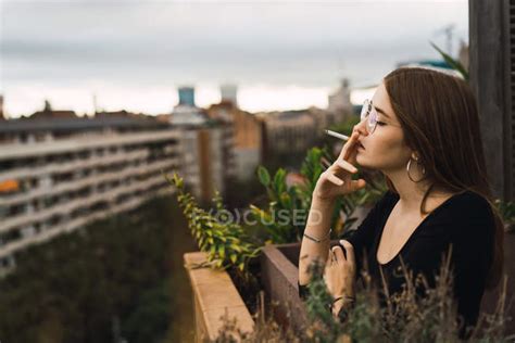 Side View Of Brunette Woman In Glasses Smoking Cigarette At Terrace