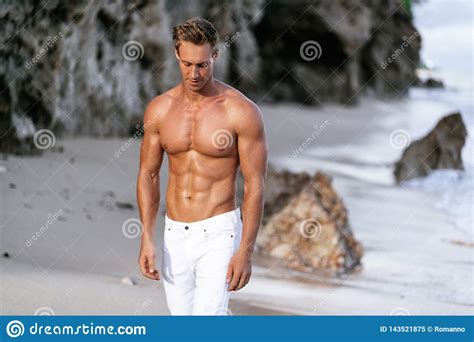 Handsome Man Shirtless And In White Pants Walking Along On Sandy Beach