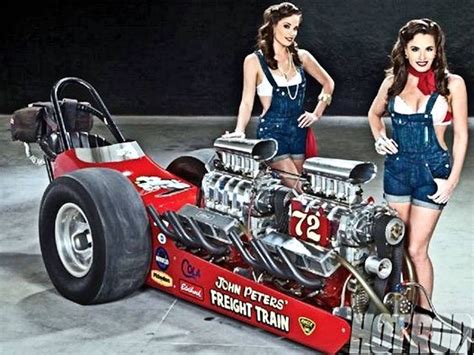 Two Women In Overalls Standing Next To A Race Car With The Engine On It