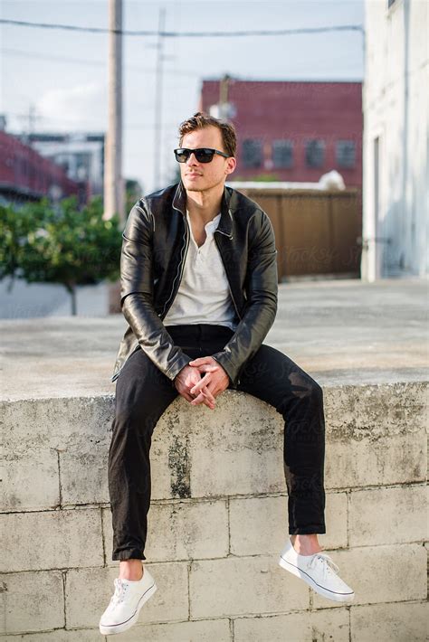 Fashionable Man In A Leather Jacket Sitting On A Concrete Wall By