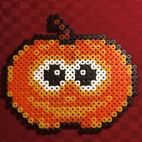 A Perler Bead Pumpkin With Black Eyes On It S Face Sitting On A Red Surface
