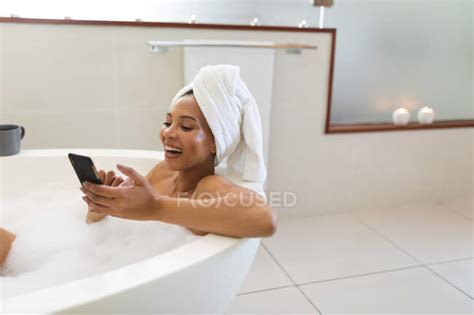 Laughing Mixed Race Woman In Bathroom Having A Bath And Using Smartphone Domestic Lifestyle