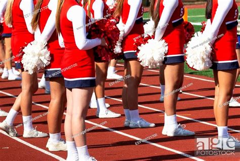 Rear View Of High School Cheerleaders Holding Pom Poms Standing On A Red Track Facing The Field
