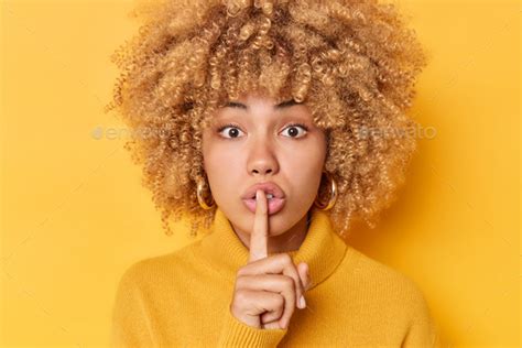 Headshot Of Surprised Curly Haired Young Woman Presses Index Finger Over Lips Makes Shush