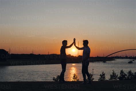 Two Friends High Fiving At Sunset Standing By The River Stock Photo