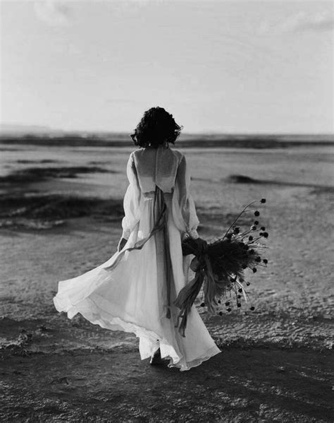 A Woman In A Long White Dress Walking On The Beach With Her Hair Pulled