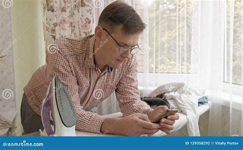 A Man Strokes His Shirt On The Ironing Board In His House And Uses A Smartphone Stock Footage