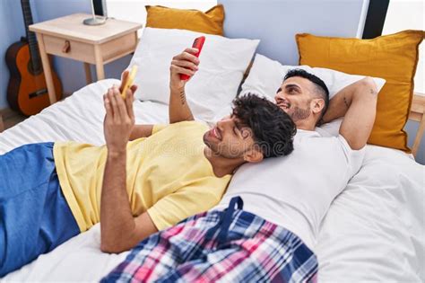 Two Man Couple Using Smartphone Lying On Bed At Bedroom Stock Photo