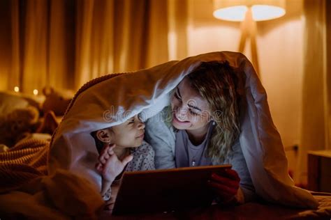 happy mother with her little daughter lying under blanket and watching movie on tablet at home