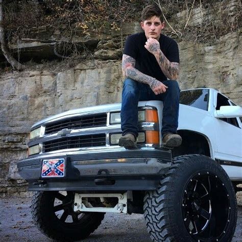 A Man With Tattoos Sitting On The Hood Of A Pickup Truck In Front Of A Cliff