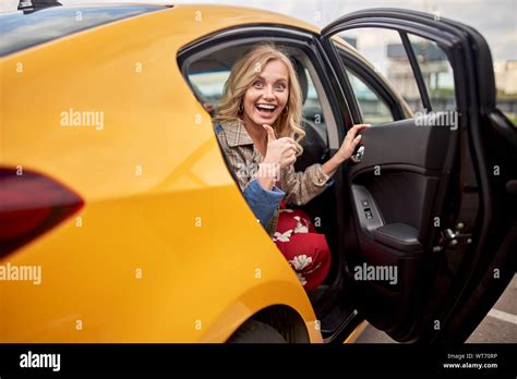 Photo Of Enthusiastic Blonde Sitting In Back Seat Of Yellow Taxi With Door Open During Day Stock