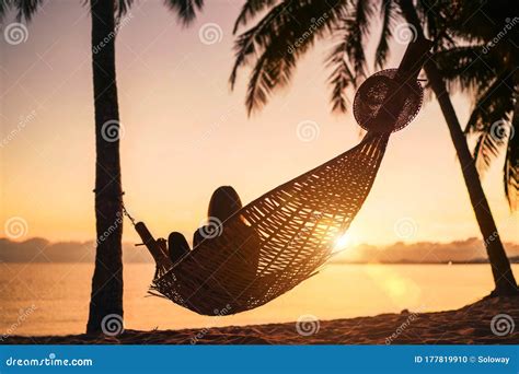 Young Woman Relaxing In Hammock Hinged Between Palm Trees On The Sand Beach At Orange Sunrise