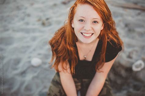 happy portrait of redhead teenage girl sitting at the beach by rob and julia campbell stocksy