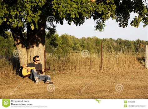 A young man in jeans, sunglasses and tshirt leaning against an old maple tree near a page wire fence playing. Guy Playing Guitar At Sunset Under Maple Tree Royalty Free ...
