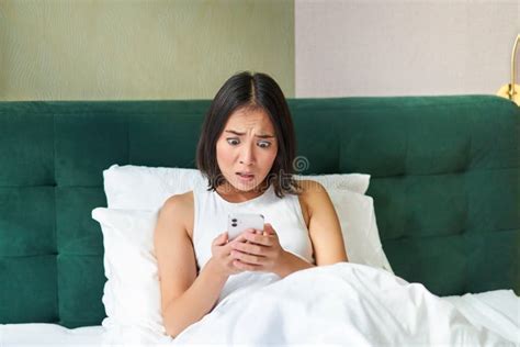 Bedroom Picture Of Asian Woman Lying In Bed Looking Scared And Shocked At Smartphone Screen