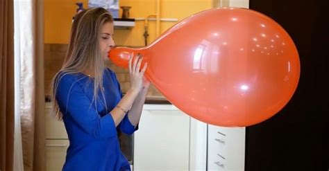 A Woman Blowing On An Orange Balloon In The Kitchen With Her Eyes Closed And Mouth Wide Open