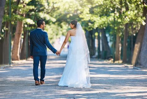 Rear View Shot Of Newlywed Couple Holding Hands And Walking Through