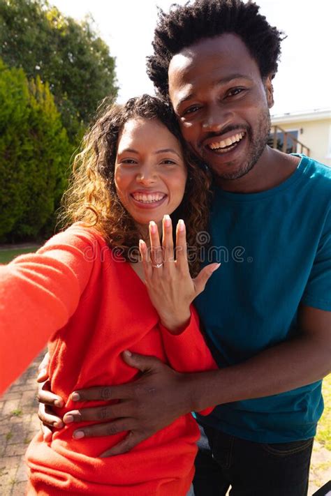 Portrait Of Smiling Young Multiracial Couple Showing Engagement Ring While Taking Selfie In