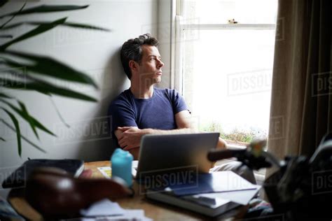 Man Sitting At Table With Open Laptop Looking Through Window Stock