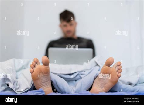 Closeup Of Bare Feet Of Man Sitting On Bed Under Blanket And Using