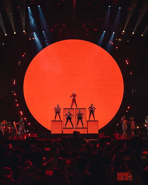 The Stage Is Lit Up With Red Lights And People Are Standing On Stools In Front Of An Orange Circle