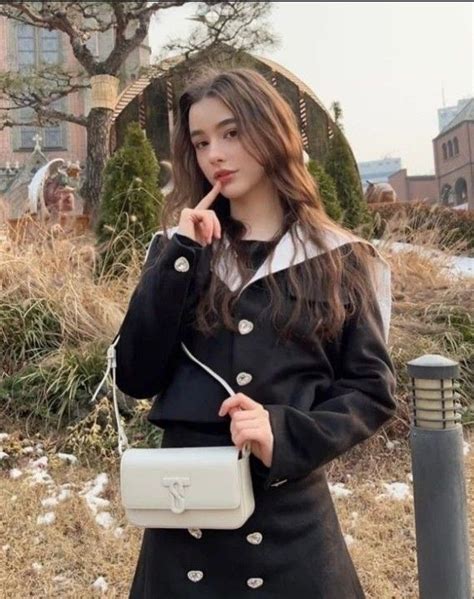 A Woman In A Black Coat Is Holding A White Purse And Posing For The Camera