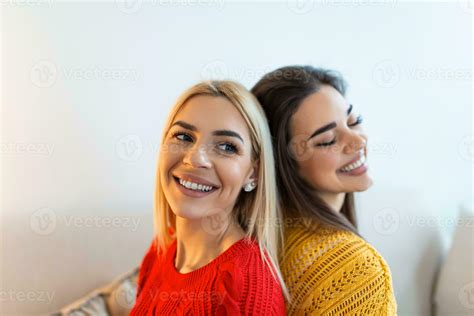 Two Beautiful Women With Blond And Brunette Hair Wearing Knitted Jumpers Sitting On The Sofa And