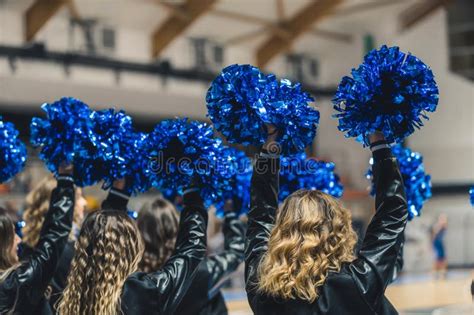 Back View Of Cheerleaders With Black Jackets Holding Blue Pom Poms Up At The Game Stock Image