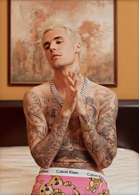 a tattooed man sitting on top of a bed with his hands clasped to his chest