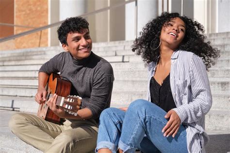 A Young Latin Couple Playing The Guitar And Smiling Sitting On Stairs