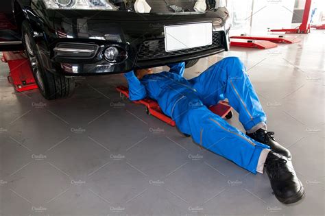 Mechanic In Blue Uniform Lying Down And Working Under Car At The Garage