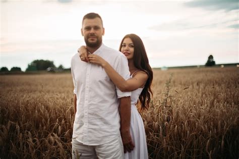 스톡 사진 밀밭에서 포옹 하는 흰 옷을 입고 그의 화려한 여자 친구를 포옹 하는 수염 난된 남자 친구의 초상화 백그라운드에서 아름 다운 밀 필드입니다 무료 사진