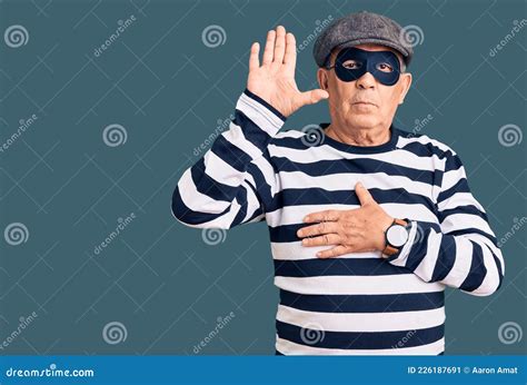 senior handsome man wearing burglar mask and t shirt swearing with hand on chest and open palm