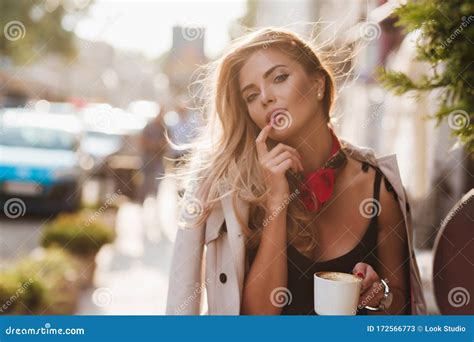 Tanned Woman With Hair Gently Waving By Wind Holding Cup Of Coffee On Blur City Background