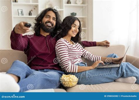 Relaxed Millennial Couple Chilling At Home Watching Tv Using Laptop