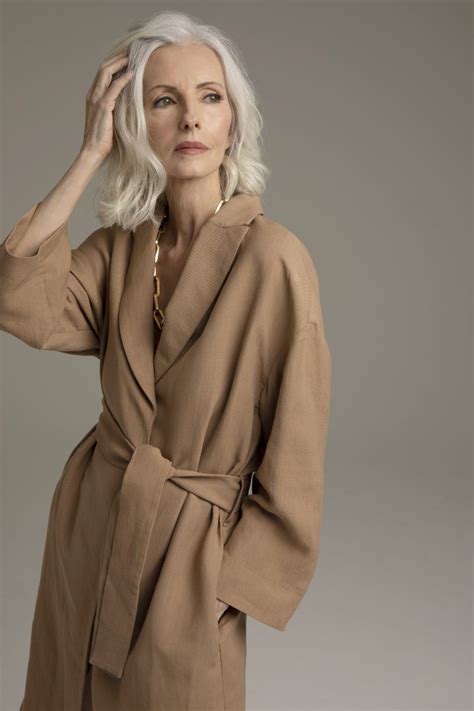 An Older Woman With White Hair Wearing A Tan Trench Coat And Gold