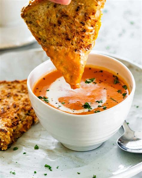 Side View Shot Of Tomato Bisque In A Soup Bowl With A A Hand Dipping Half A Grilled Cheese In
