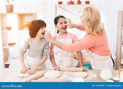 Attractive Grandmother Smeared With Flour Nose Of A Joyful Grandson And Granddaughter In Kitchen