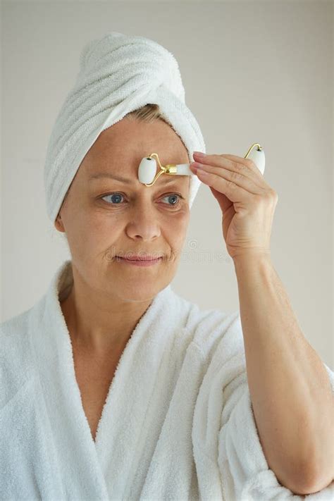 Close Up Of Mature Woman In White Bathrobe With Towel On Head Doing Facial Massage With Quartz