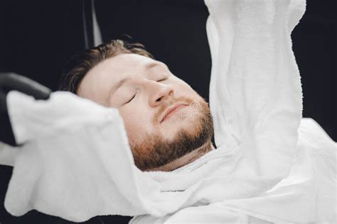 man sits in barbershop chair and steam his face with hot towel in front of royal beard shaving