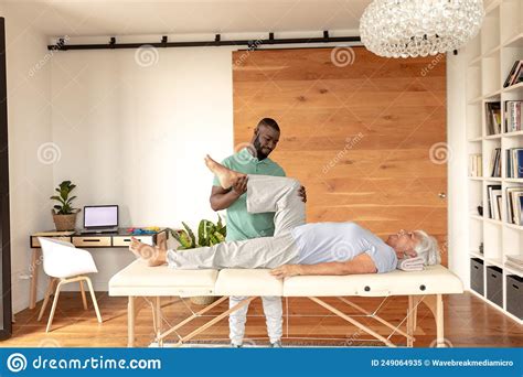 African American Male Physiotherapist Giving Leg Massage Therapy To Caucasian Senior Woman At