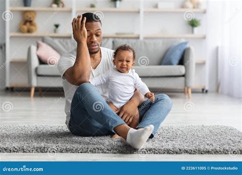 Exhausted Black Father Sitting With Crying Newborn Baby On Floor At
